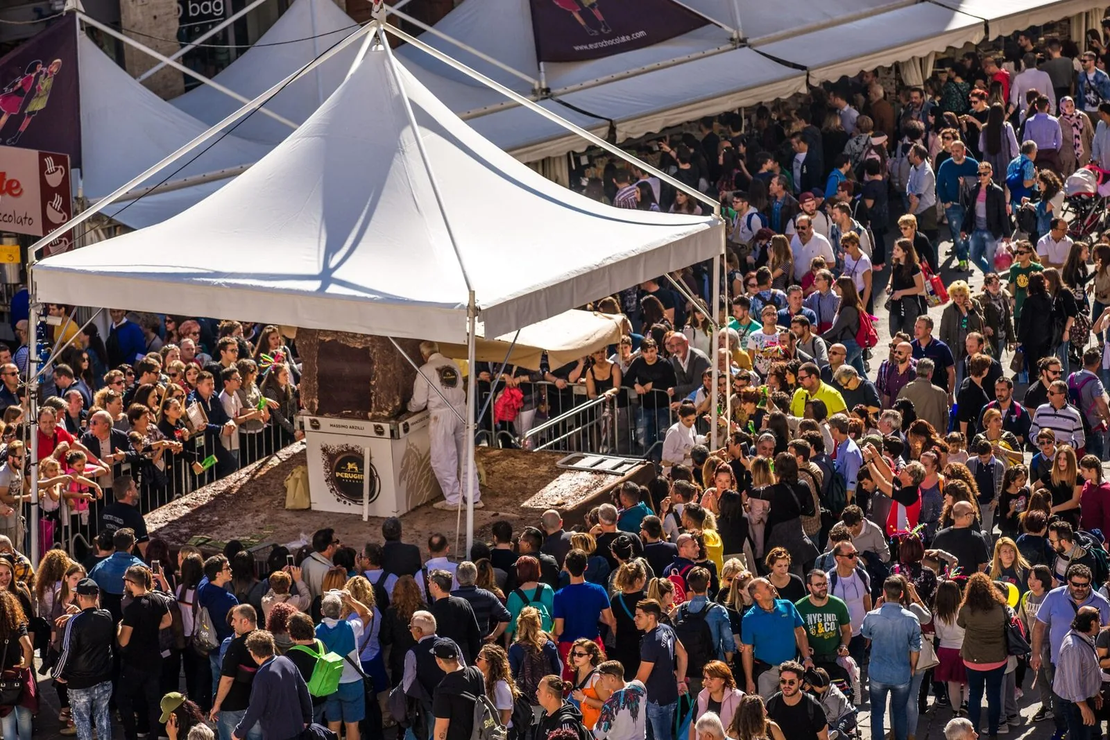Scultori al lavoro su un grande blocco di cioccolato sotto un gazebo bianco, circondati dal pubblico di Eurochocolate a Perugia